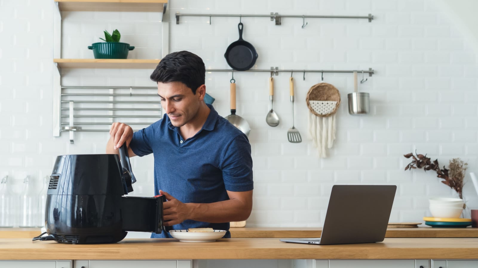 Happy handsome man cooking healthy food with recipe by Air Fryer machine and using laptop computer in the modern kitchen at home on sunny day. Watching or Learning with Technology