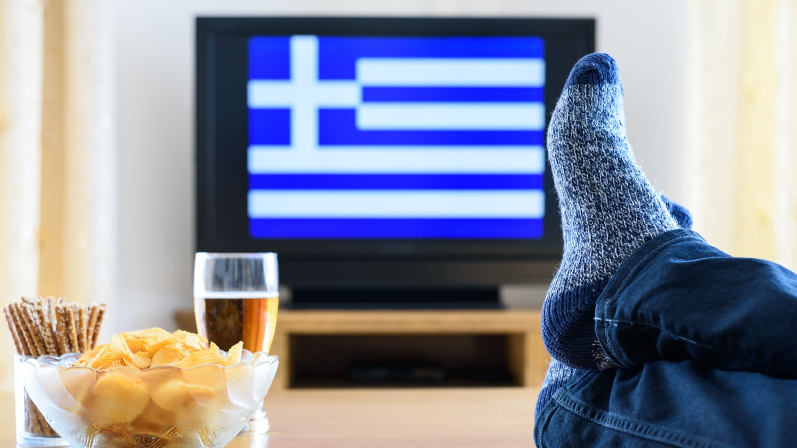 man watching Greece flag on TV screen with legs on table - stock photo