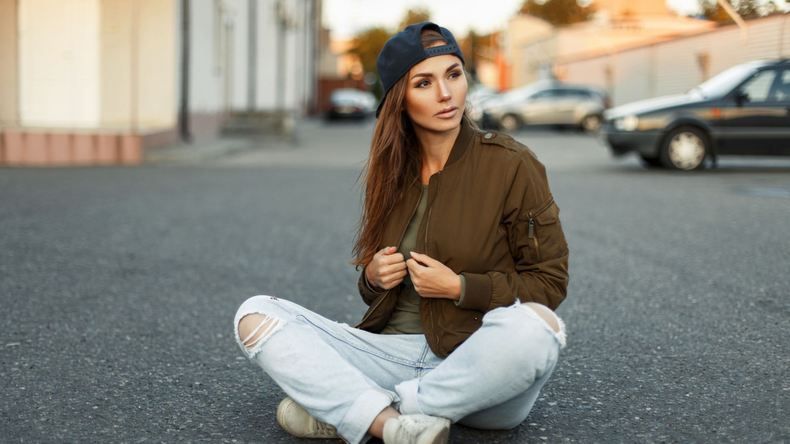 Beautiful young woman with freckles in a fashionable jacket and blue ragged jeans sits on the asphalt