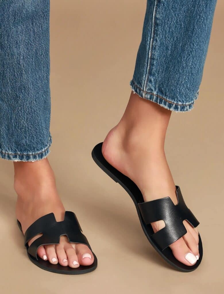 A model wears a pair of Cadence Black Vachetta Leather Slide Sandals - a solid Hermès Oran sandals dupe in black leather.