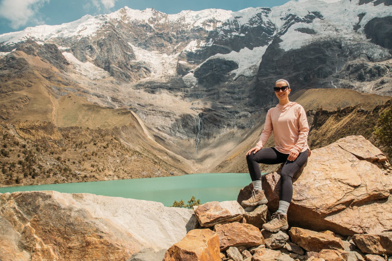 Lindsey of Have Clothes, Will Travel wearing earth leggings and a pink REI shirt sitting on a rock by a bright blue colored lake in Peru