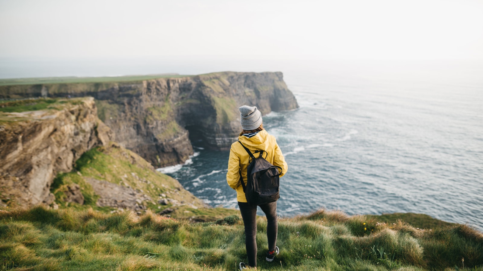 Young backpacker girl standing on the Cliffs of Moher in yellow jacket with backpack