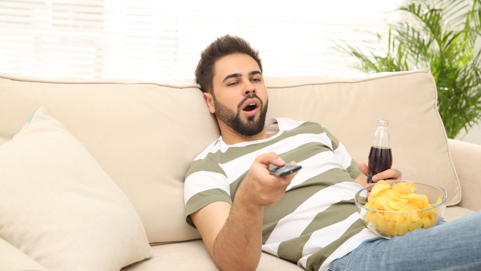Lazy young man with chips and drink watching TV on sofa at home