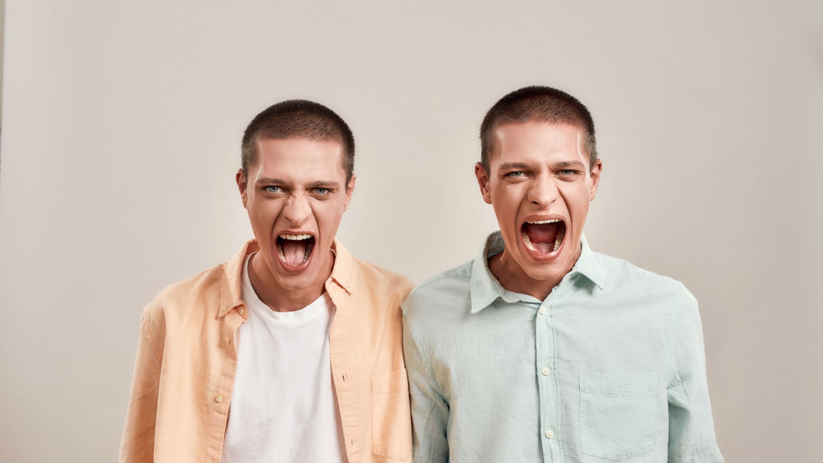 Close up portrait of two young angry twin brothers shouting and looking at camera while posing together isolated over beige background. Family relationships, human emotions