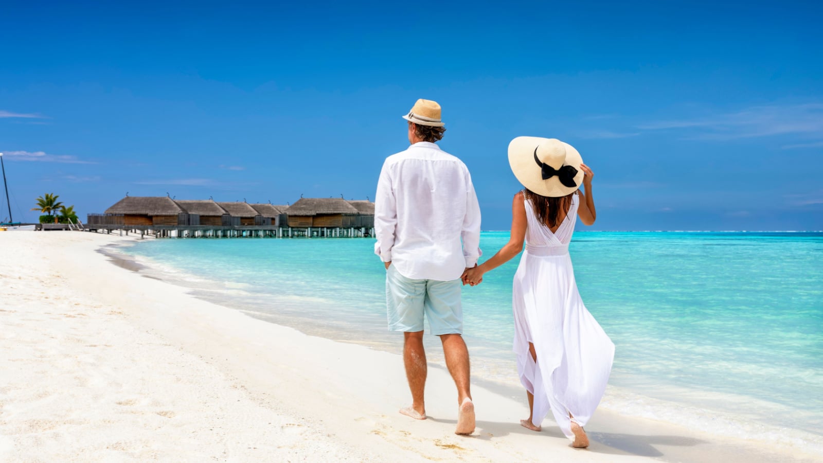 Happy couple in white clothing and with hats walks down a tropical beach with turquoise sea in the Maldives islands