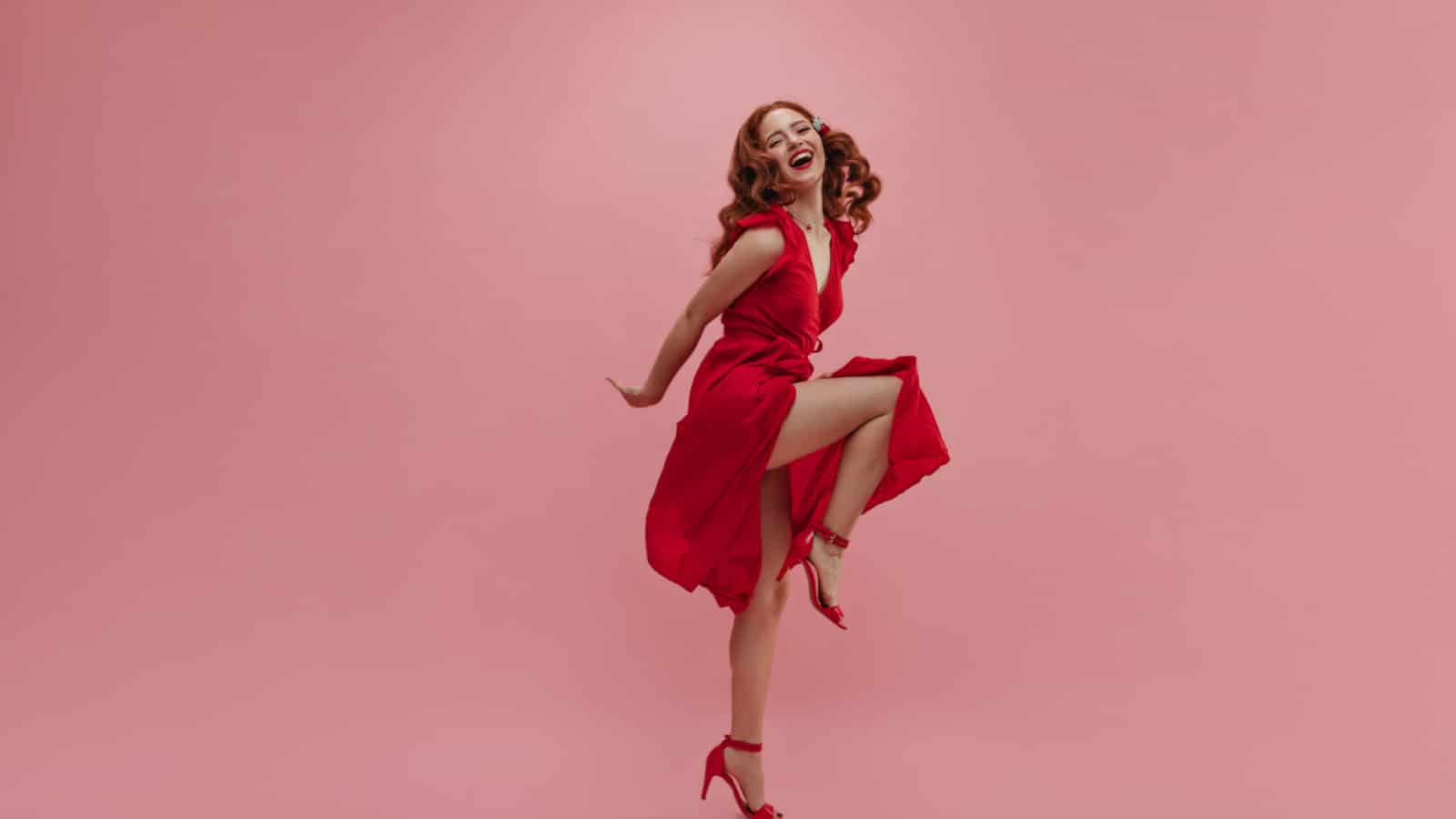 In full growth young red-haired woman dances vigorously for photo in studio. Beauty in red dress with slit and bare leg in high-heeled shoes. Leisure concept of youth, active lifestyle