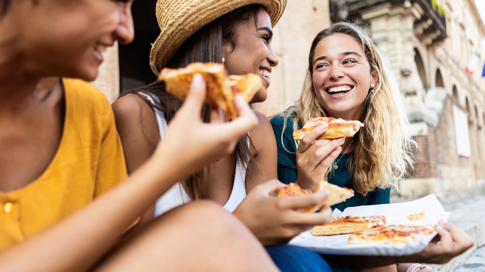 Three young multiracial women laughing while eating a piece of pizza in italian city street - Happy female friends enjoying holidays together in Italy - Friendship, travel and tourism concept