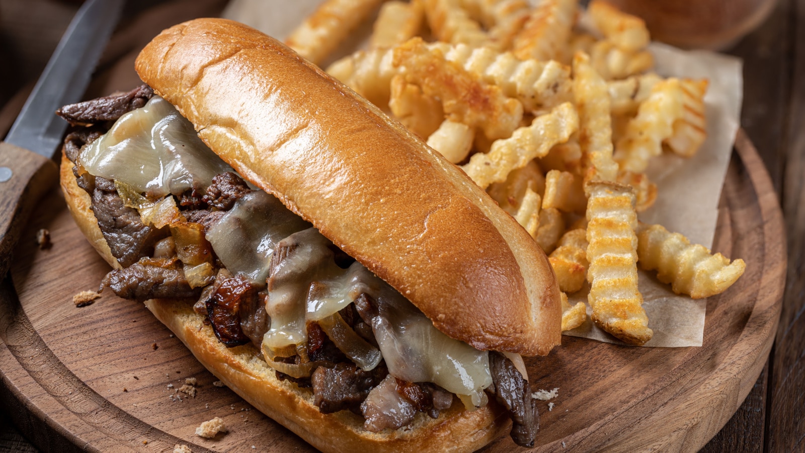 Philly cheesesteak sandwich made with steak, cheese and onions on a hoagie roll with french fries on a wooden board