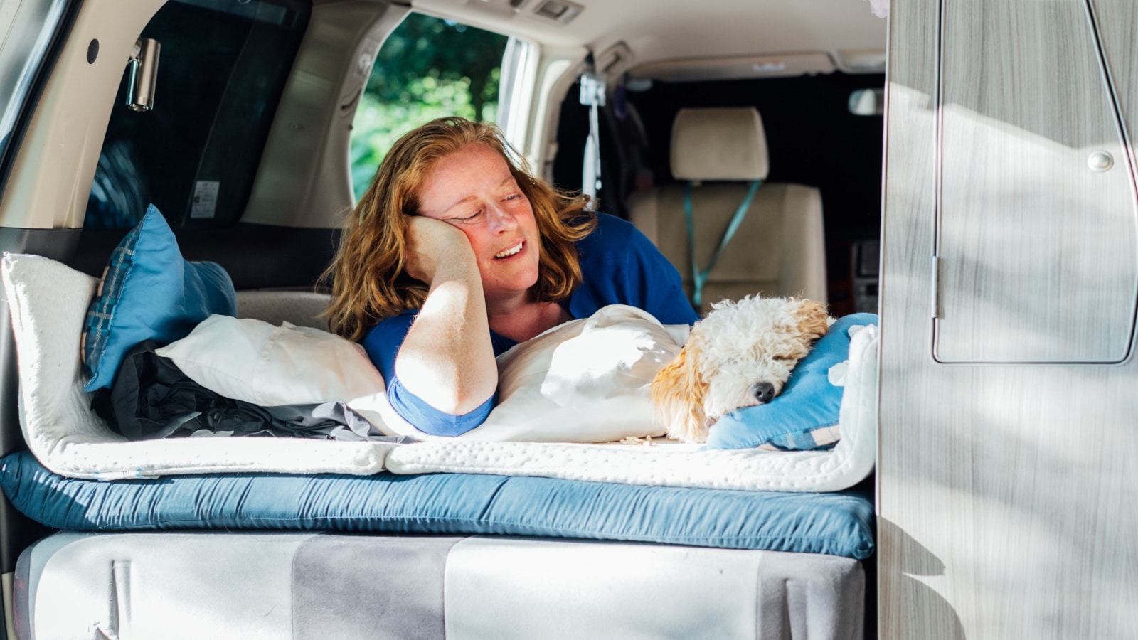 Smiling middle-aged woman looking on sleeping cockapoo puppy pet while lying in camper car vehicle during road trip. Window view. Best friend. Enjoying free lifestyle, travel, vacation, freedom.