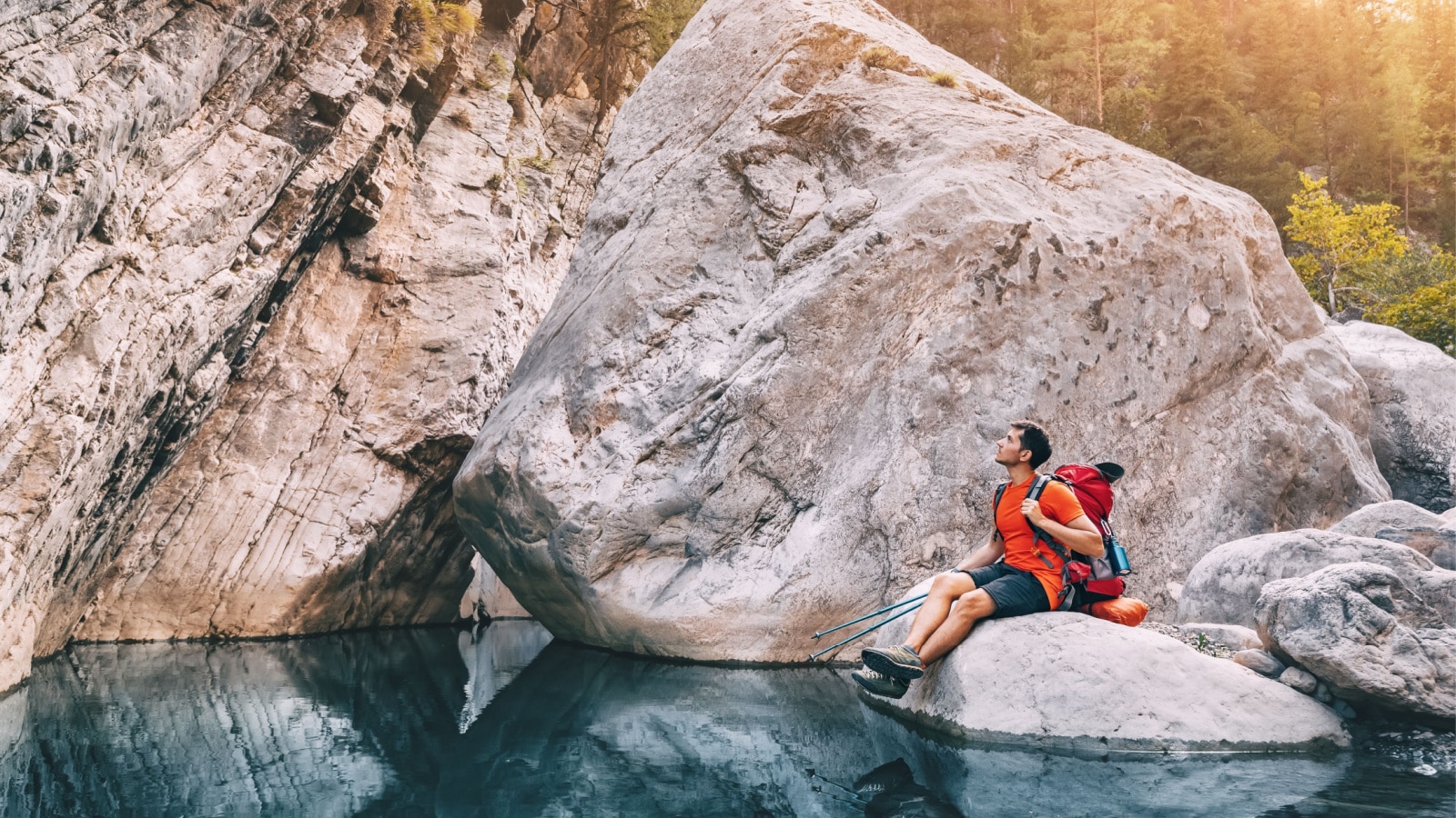 Happy hiker man with trekking backpack on a trail in mountains on a bank of a scenic river stream in the deep canyon or gorge with huge rocks and stones