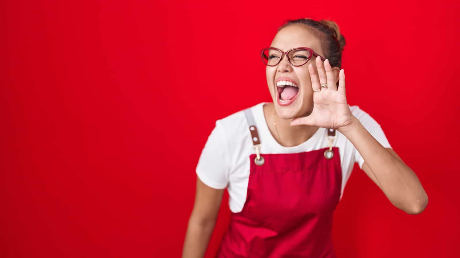 Young hispanic woman wearing waitress apron over red background shouting and screaming loud to side with hand on mouth. communication concept.