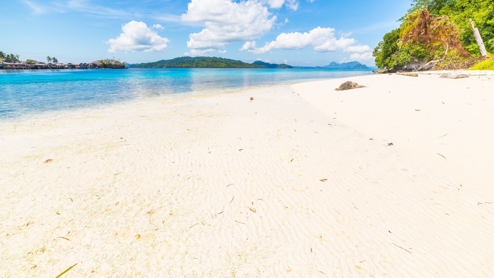 White sandy beach, turquoise transparent water and lush green jungle in the remote Togean (or Togian) Islands, Sulawesi, Indonesia. Islets and fishermen village in the background. wide angle view.