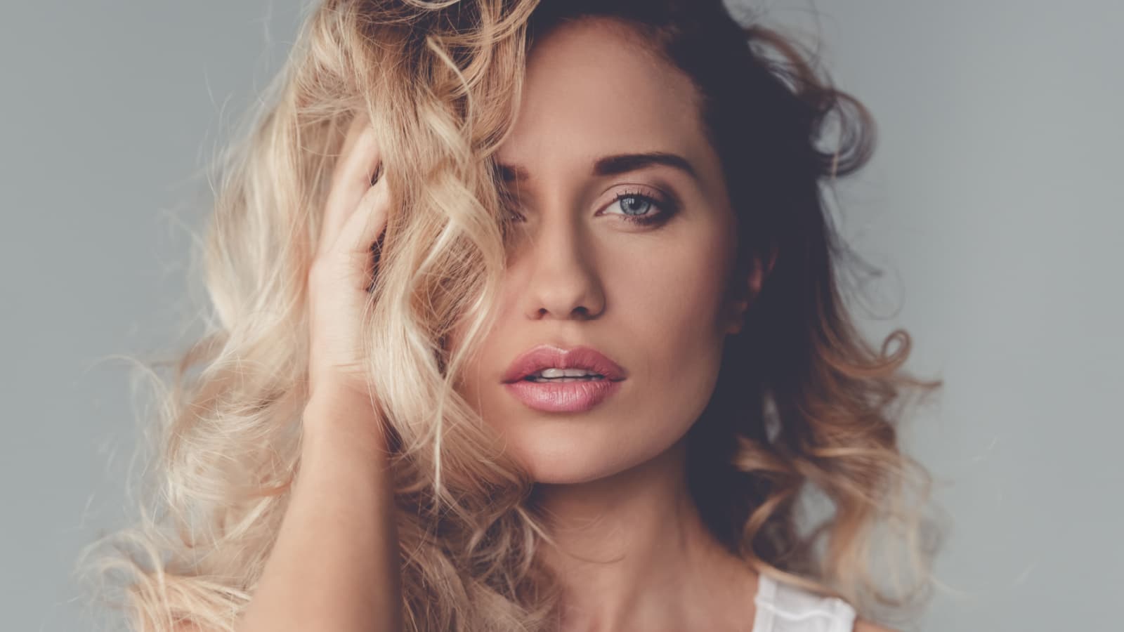 Portrait of beautiful blonde girl touching her hair and looking at camera, on gray background