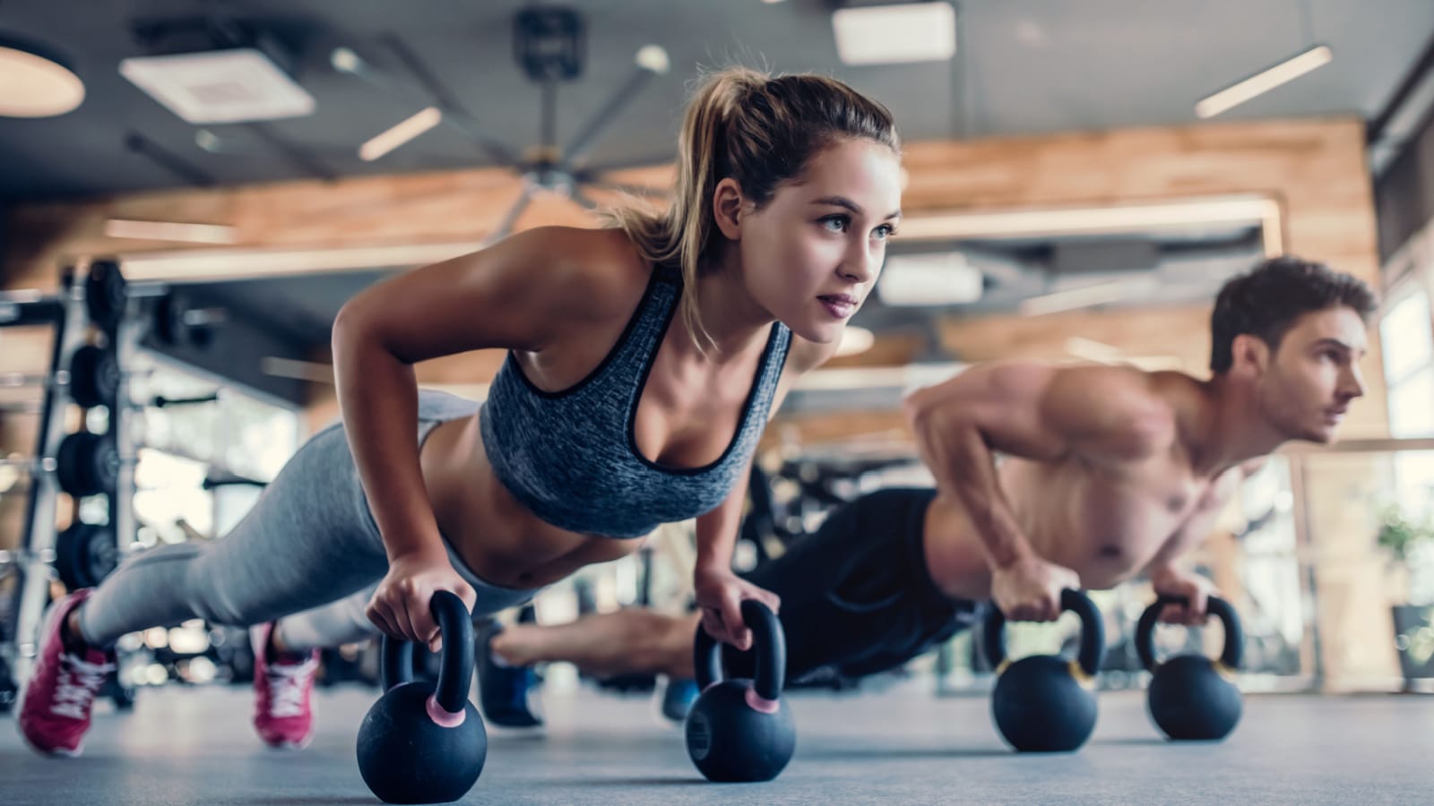 Young couple is working out at gym. Attractive woman and handsome muscular man are training in light modern gym. Doing plank on kettlebell. Push-up on weights.