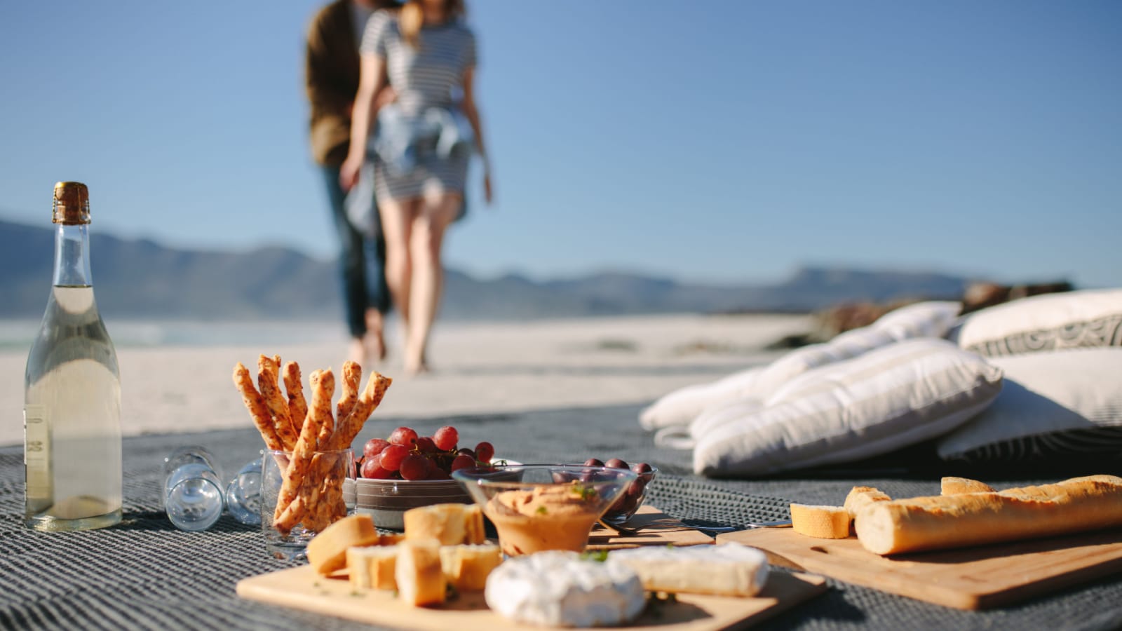 Food and drink on the blanket at the beach with couple walking towards it. Picnic setting on the beach with man and woman in background.