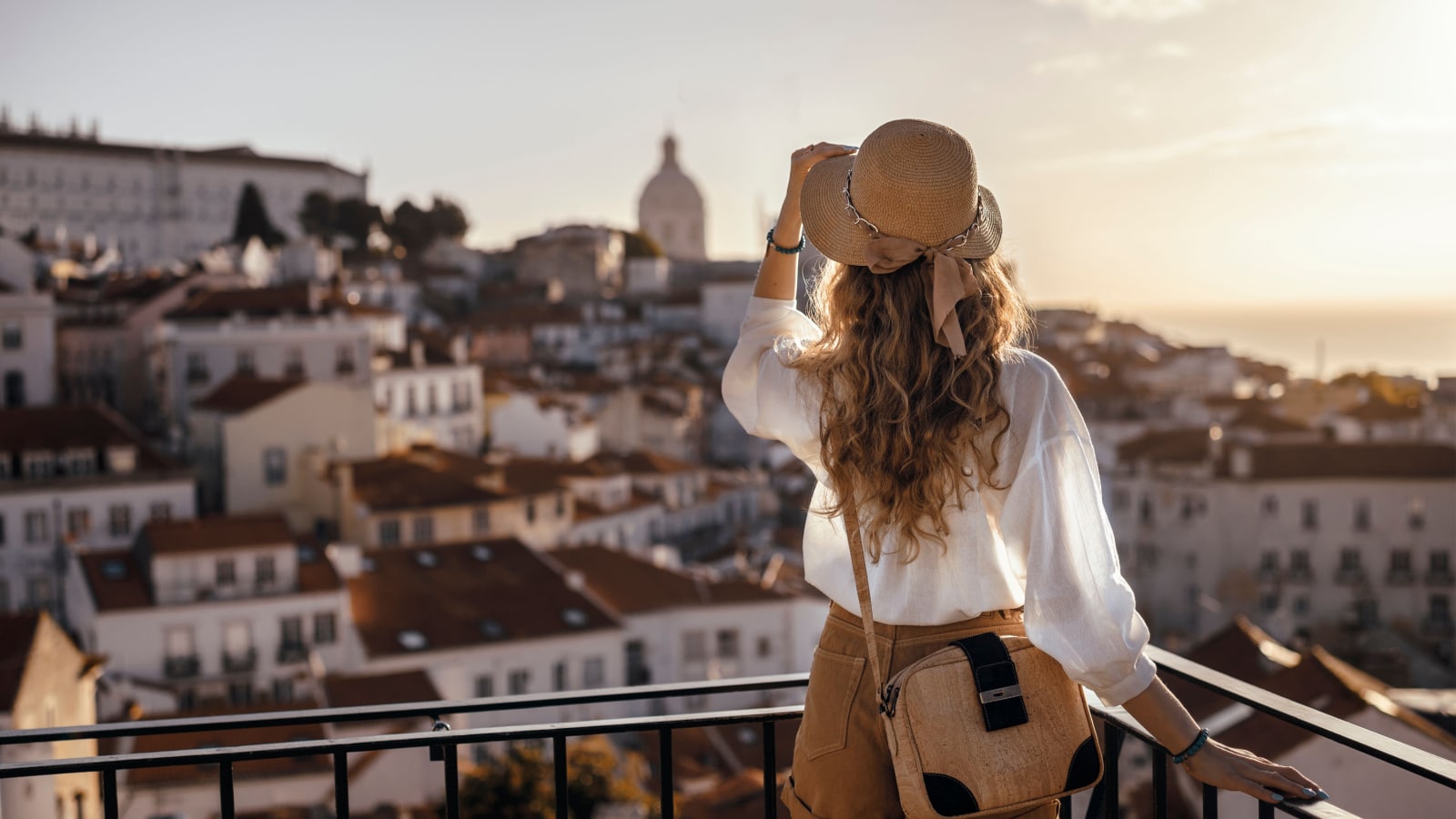 Blonde woman standing on the balcony and looking at coast view of the southern european city with sea during the sunset, wearing hat, cork bag, safari shorts and white shirt
