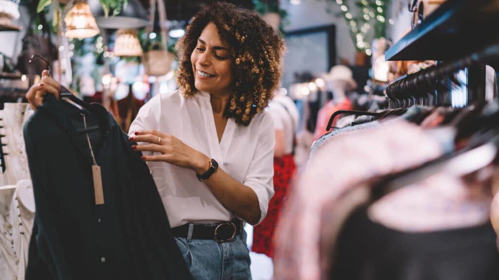 Smiling female standing near hangers with brand wear and laughing during Black Friday shopping with discounts, funny hipster girl enjoying pastime for update her wardrobe spending day in boutique