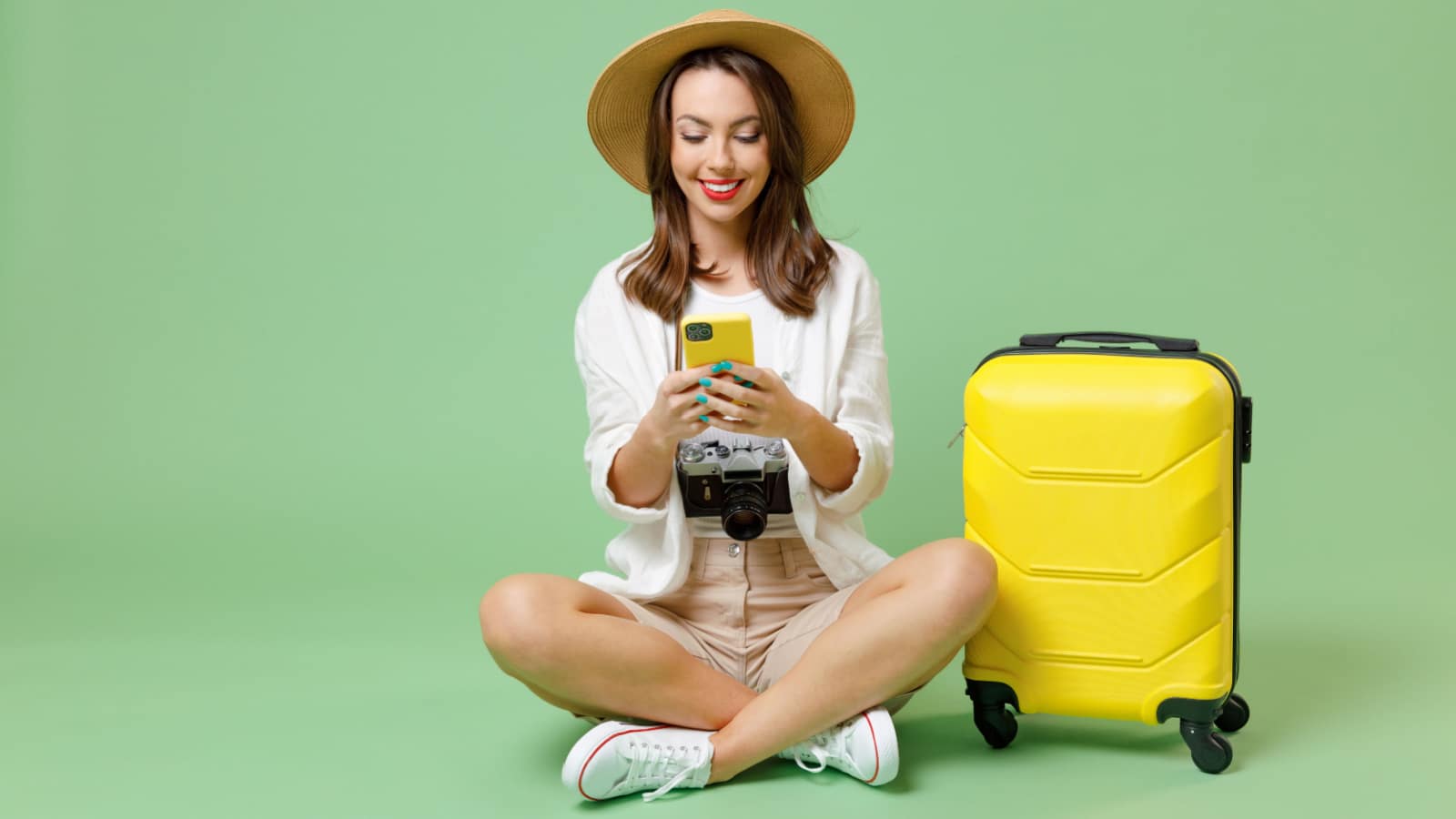 Full length traveler fun tourist woman in casual clothes hat hold suitcase using mobile cell phone sit isolated on green background Passenger travel abroad weekends getaway Air flight journey concept