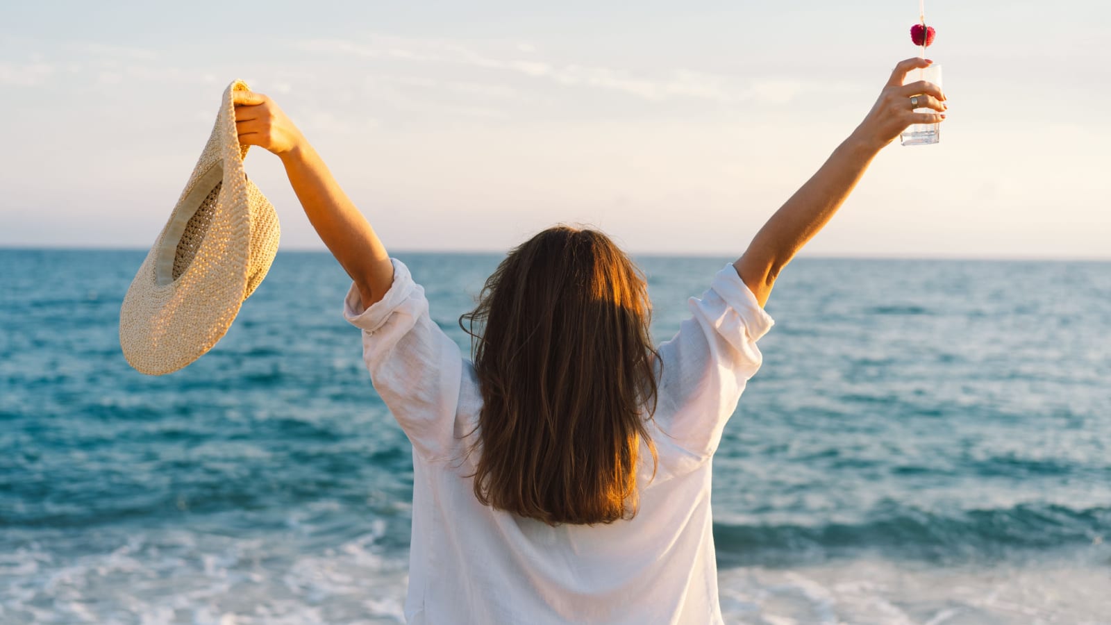 Portrait of a happy young girl in a straw hat with a cocktail in hand on a background of beautiful sea. Woman from behind. The girl looks at the magical sea. Freedom and happiness