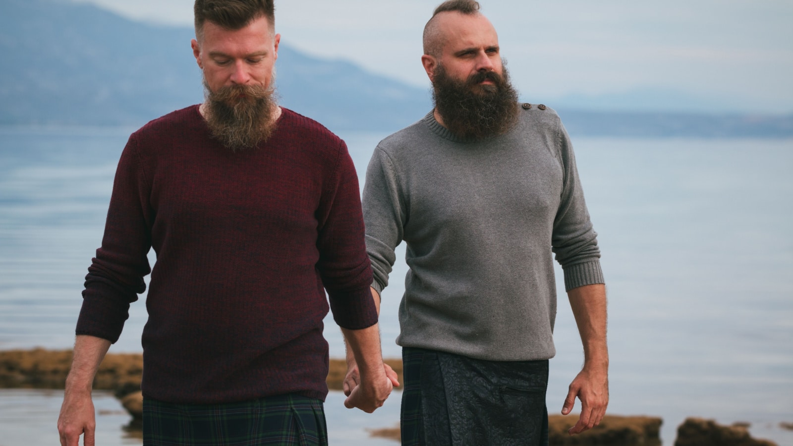 Fashionable gay couple in kilts holding hands while walking by sea side. Long beard male on the beach, serious faces, overcast day.