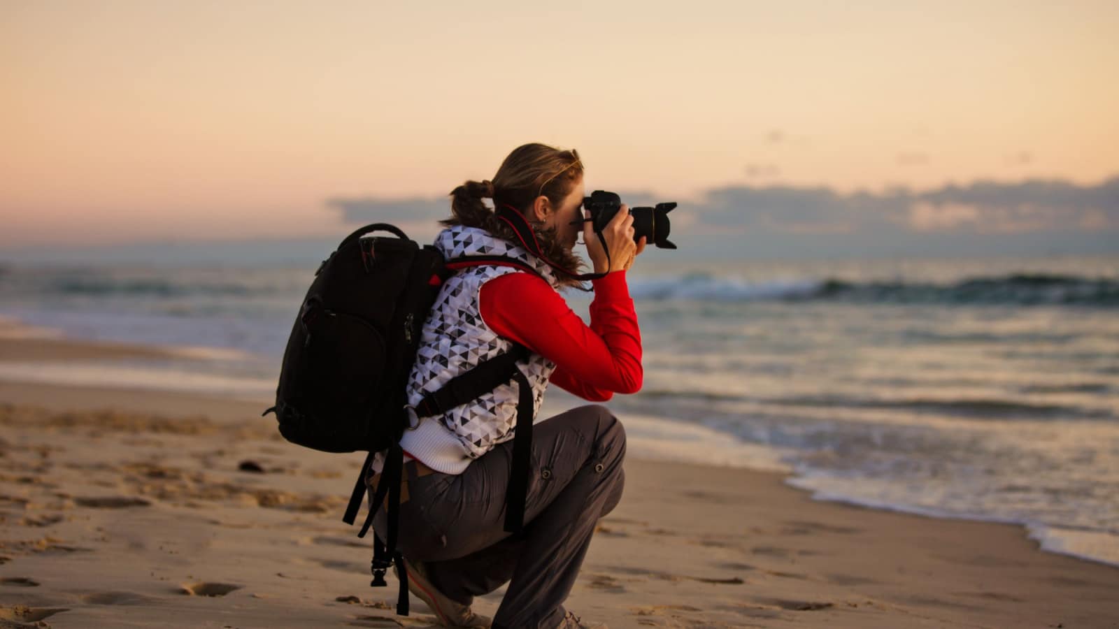 Young woman photographer with backpack taking pictures with SLR camera at sunset on the winter beach.