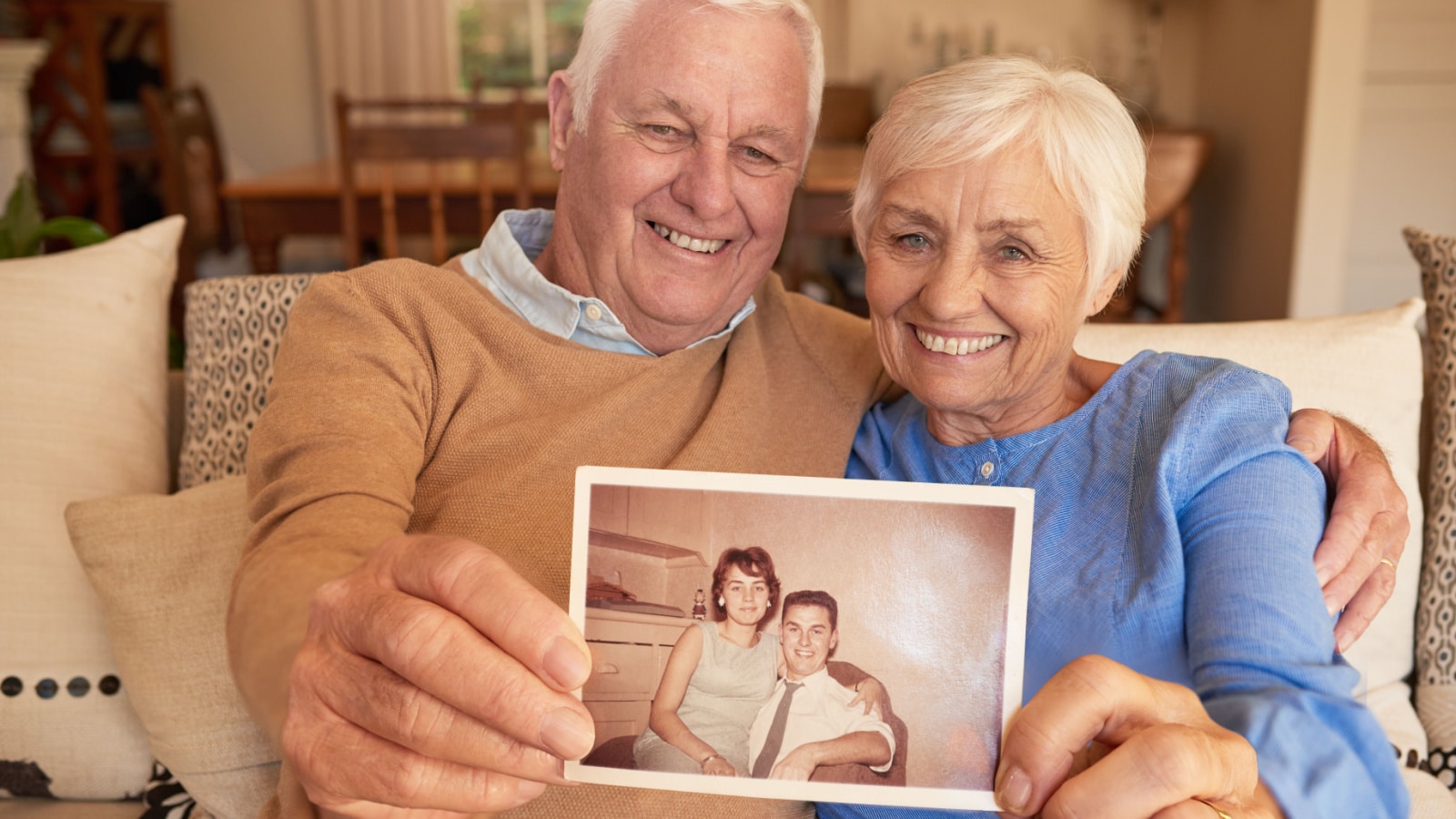 Portrait of a smiling senior couple holding up an old photograph of themselves when they were young while sitting on their sofa at home