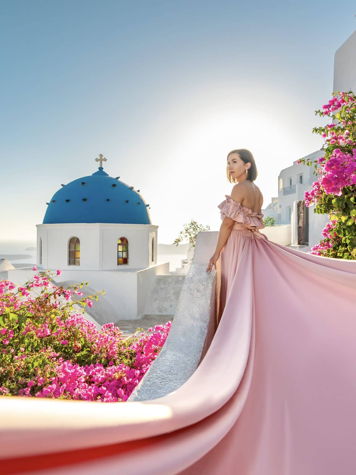 Lindsey of Have Clothes, Will Travel wearing a pink dress with a blue church dome and pink flowers in the background in Imerovigli, Santorini Greece