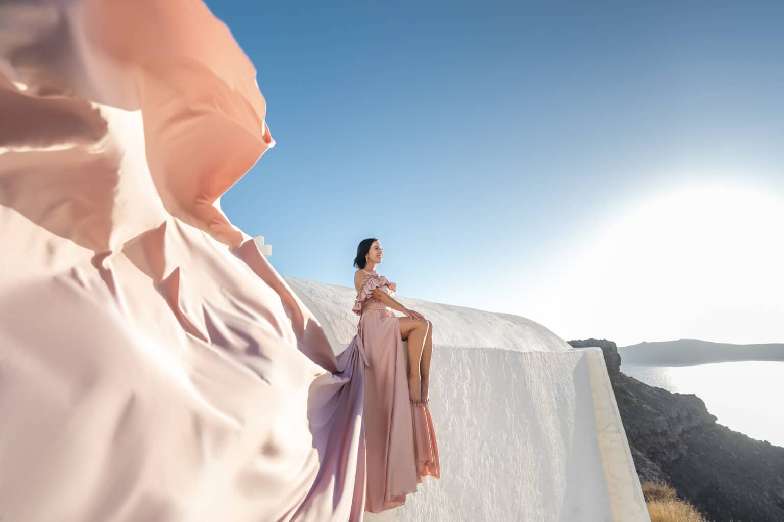 Lindsey Puls of Have Clothes, Will Travel sitting on a white ledge with a pink flying dress in Santorini Greece