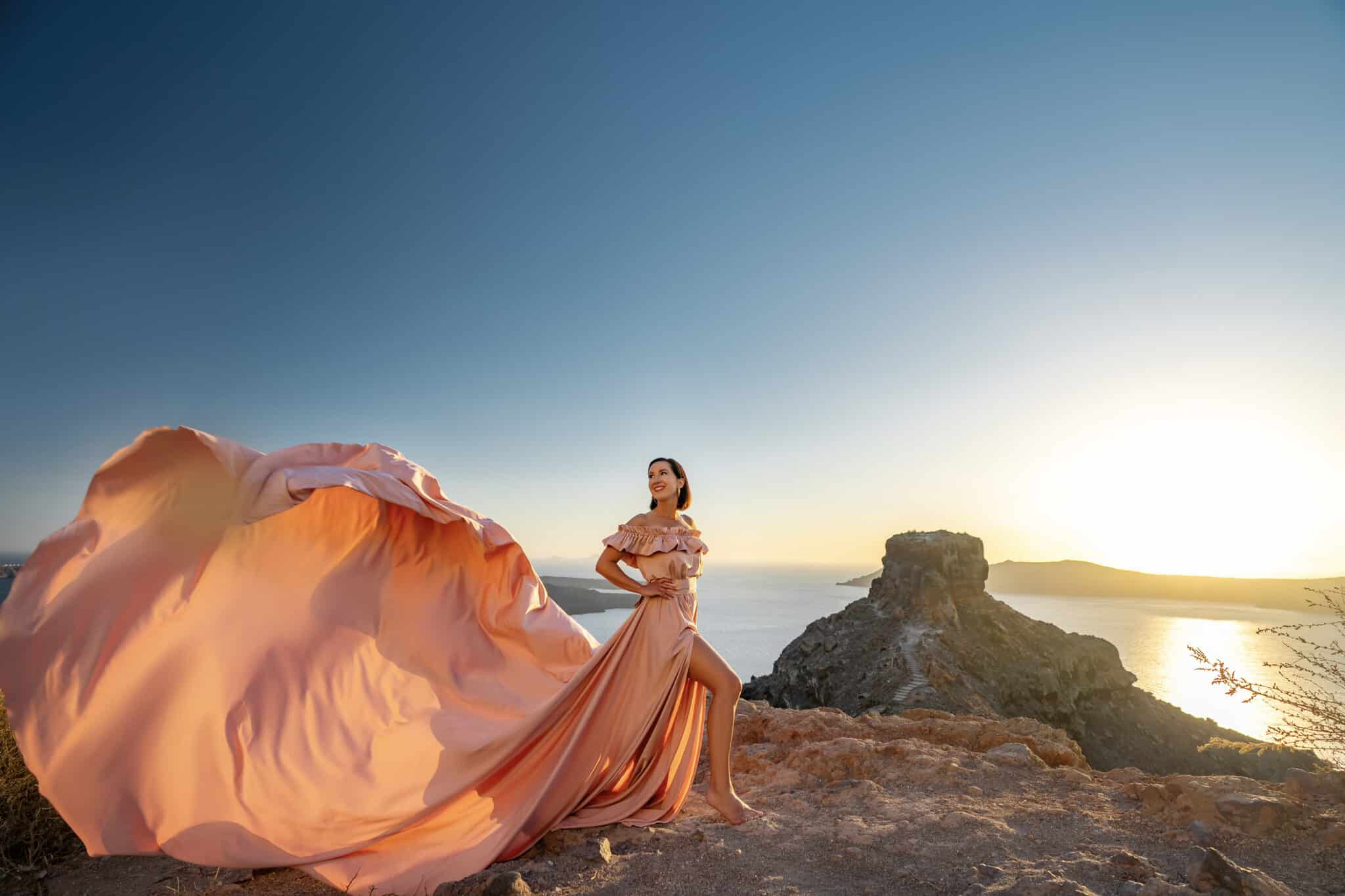 Lindsey Puls of Have Clothes, Will Travel standing on a cliff at sunset with a pink flying dress in Santorini Greece