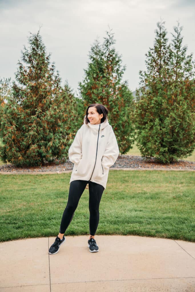 Lindsey wearing a white oversized Nike hoodie that zips up, black leggings and black shoes. She's standing on a concrete driveway, in front of a green lawn with tall pine trees behind her.