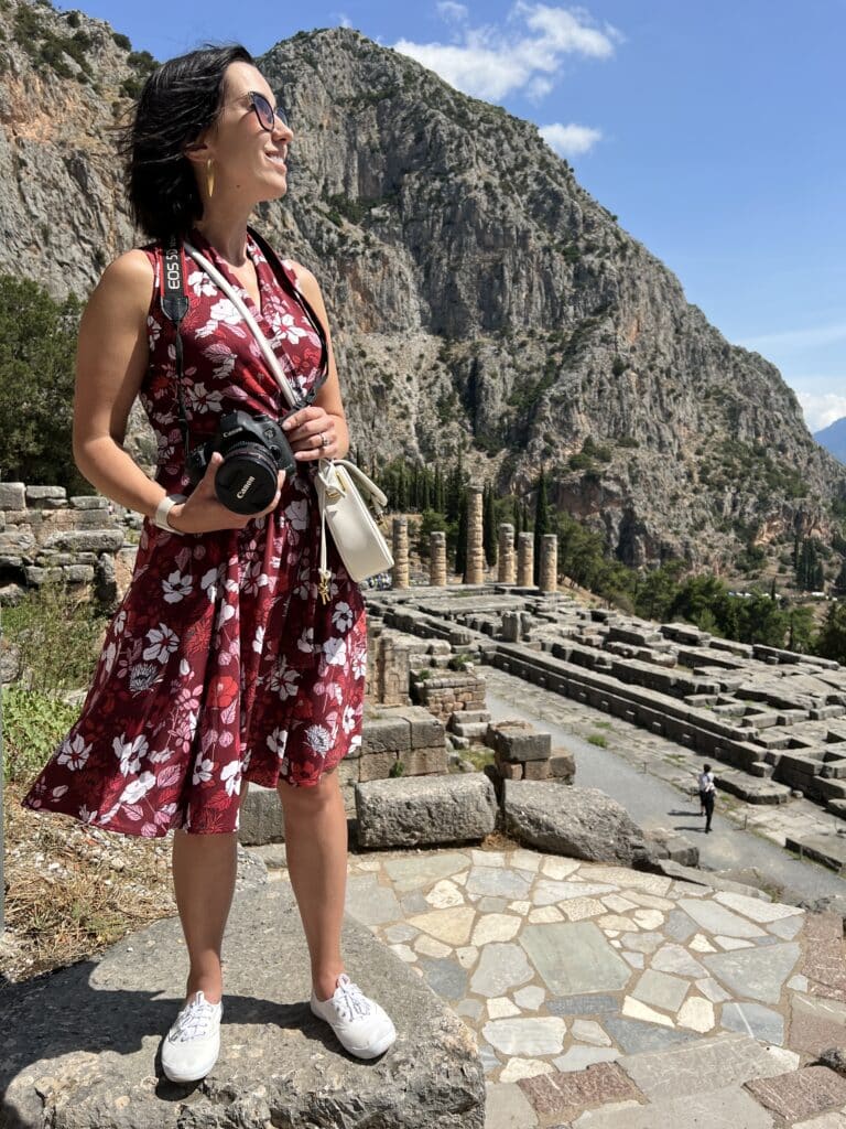 Lindsey wearing a floral Karina dress and white keds. She's standing outside on a paved stone pathway, admiring the scenery in Delphi Greece.