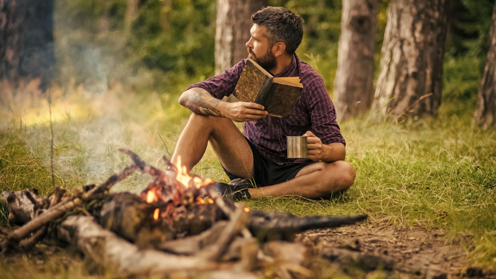 Summer vacation, activity. Man traveler read and drink at campfire flame. Camping, hiking, lifestyle. Hipster hiker with book and mug at bonfire in forest. Sustainable education, environment concept.