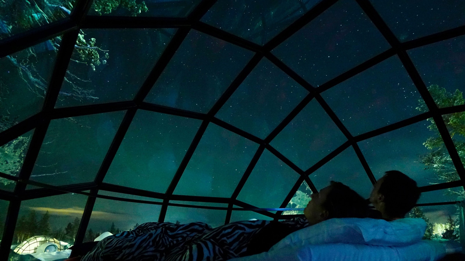 CLOSE UP Young Caucasian tourist couple observing the night sky from a cool glass house in the Scandinavian wilderness. Girlfriend and boyfriend enjoying a romantic evening in a cool glassy igloo.