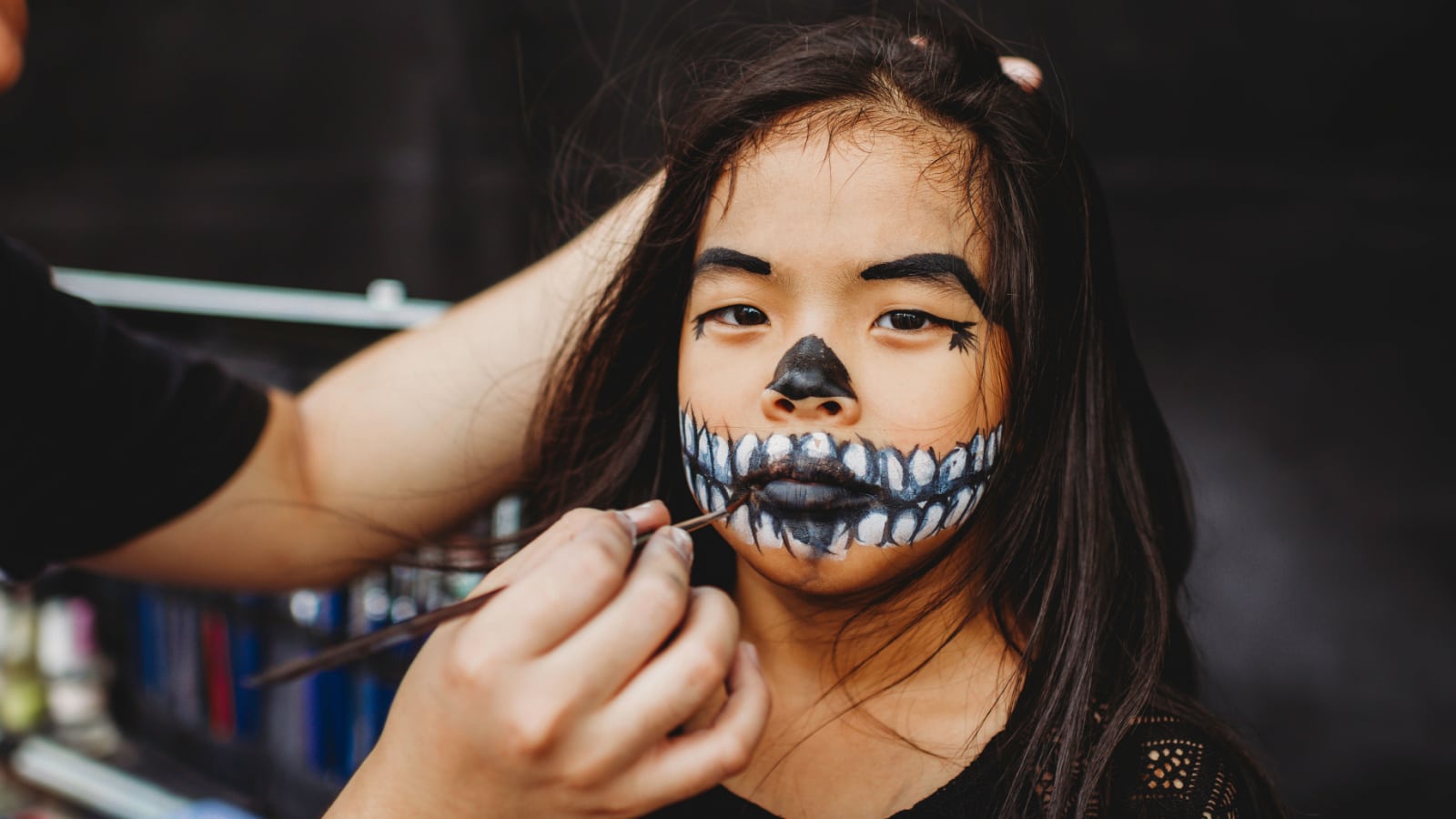 Young Asian Chinese girl with skeleton face make up. Make-up artist holding brush to paint mouth area. Skeleton design for chinese girl in Asia. Outdoor studio in the street. Drama competition.