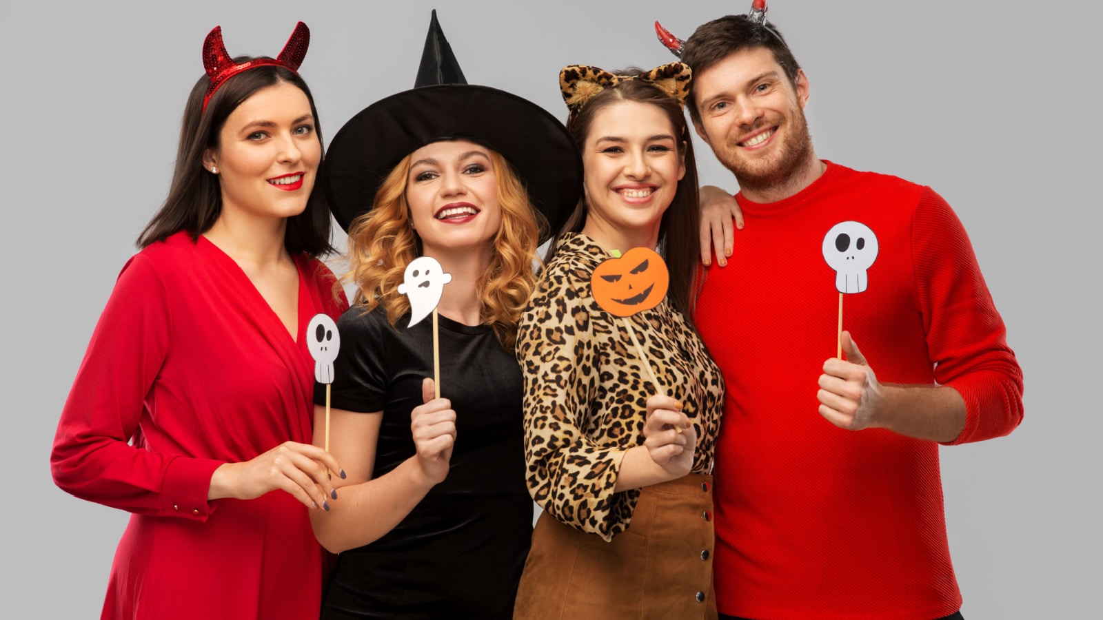 friendship, holiday and photo booth concept - group of happy smiling friends in halloween costumes of devil, witch and cheetah with party props over grey background