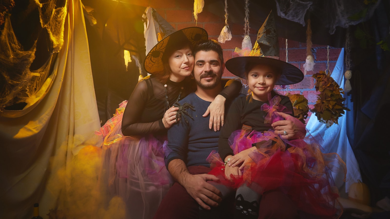 Mom, dad and daughter in costumes for holiday of halloween in dark decoration and with nice yellow light. Funny nice family during carnival in room. Mother, father and girl during funny photo shoot