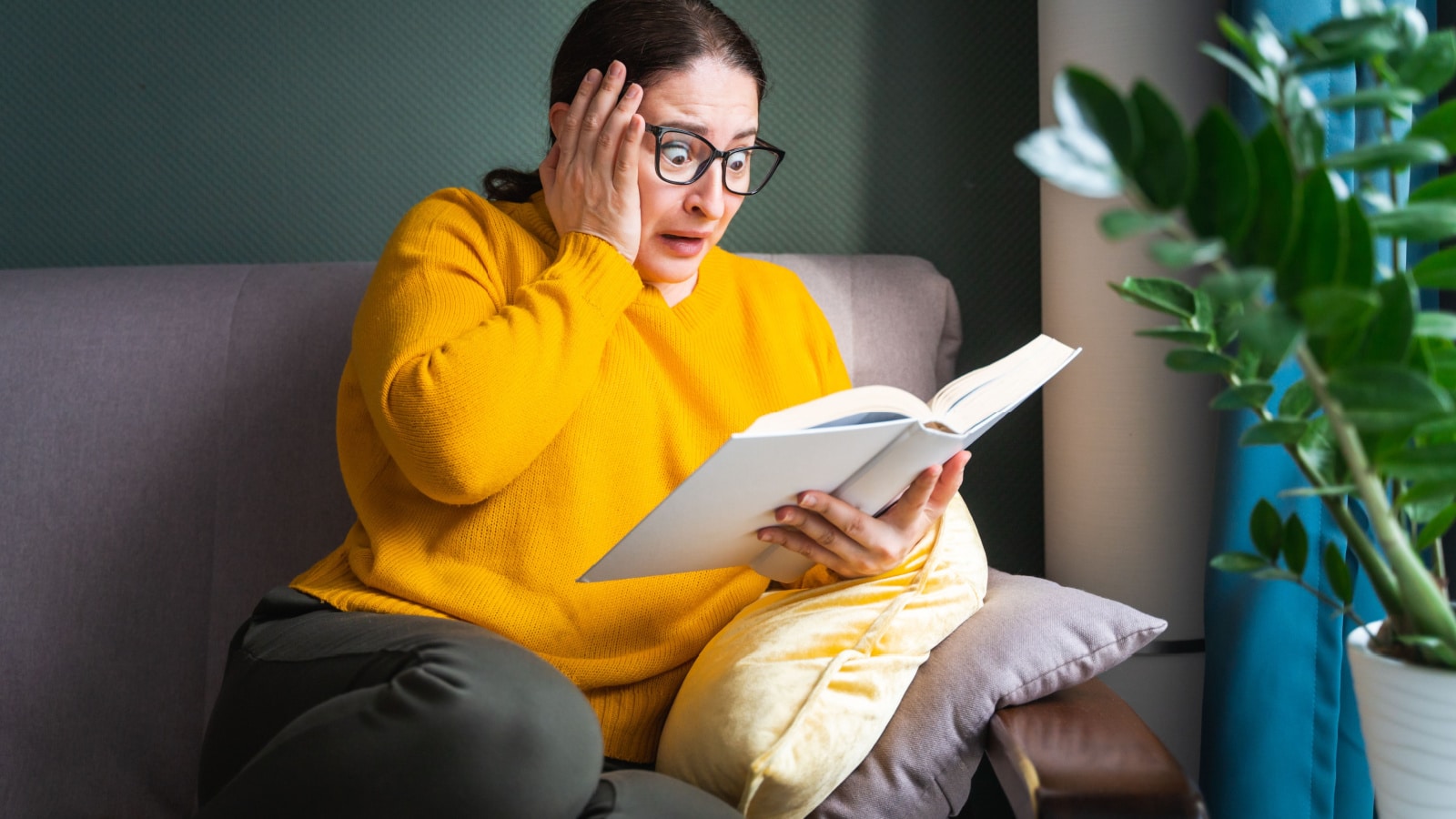 Frightened scared middle-aged woman in eyeglasses reading horror novel, thriller or scary stories while sitting on sofa at home, emotional shocked female with book in hands feeling terrified