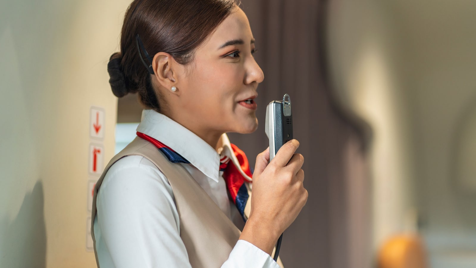 Asian female flight attendant holds a microphone talking to passengers on board an airliner serving passengers during their journey.