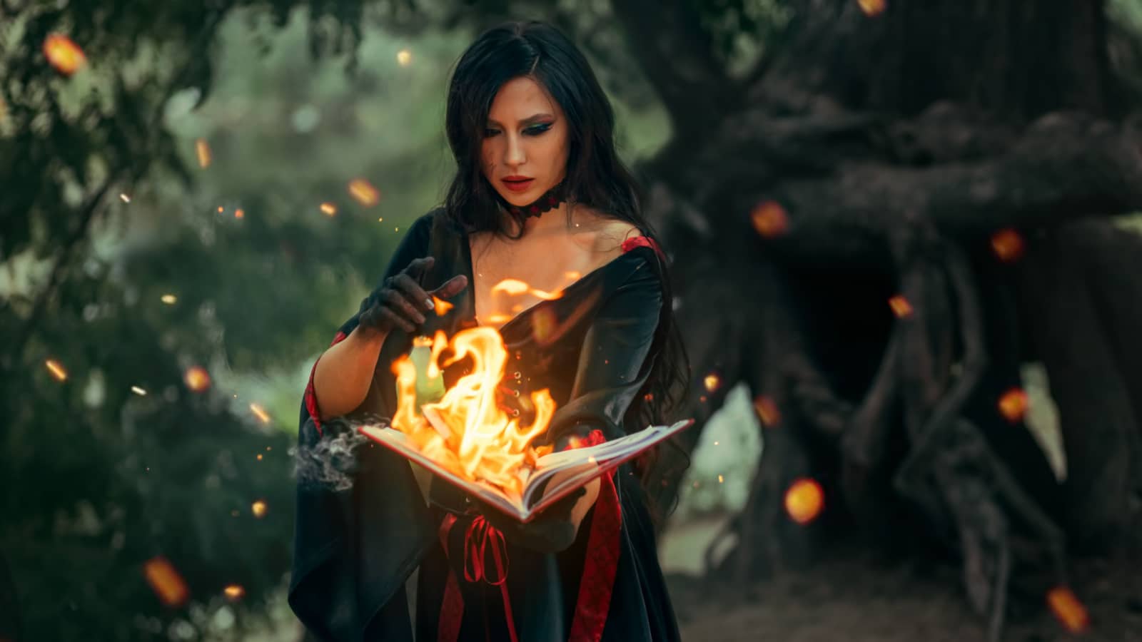 Fantasy halloween woman witch holds old burning magic book in hand, reads spell Paper page in bright flame fire light. Gothic girl sexy face in ashes, black dress costume. Dark night forest trees. Art