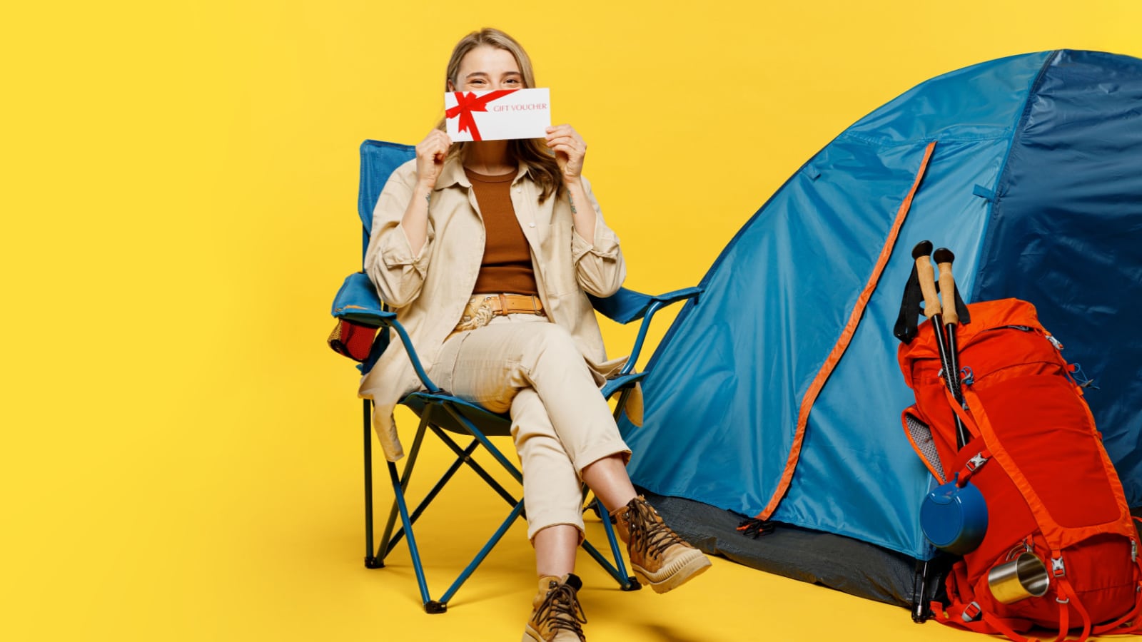 Full body young woman sit near bag with stuff tent hold gift coupon voucher isolated on plain yellow background. Tourist leads active lifestyle walk on spare time. Hiking trek rest travel trip concept