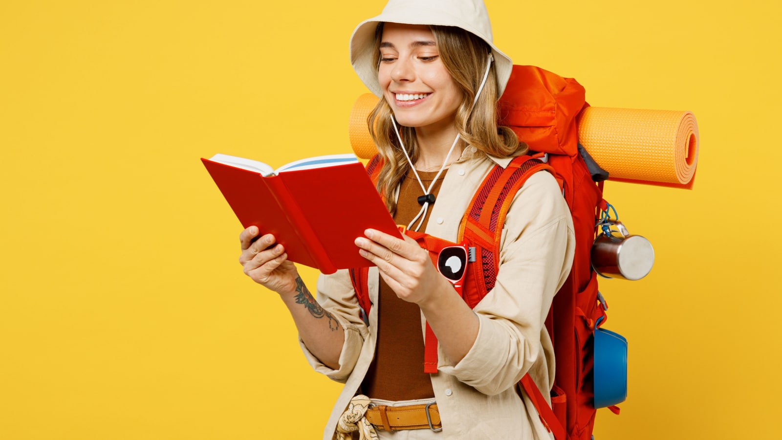 Young smiling happy fun woman carry backpack with stuff mat read book diary isolated on plain yellow background. Tourist leads active lifestyle walk on spare time. Hiking trek rest travel trip concept