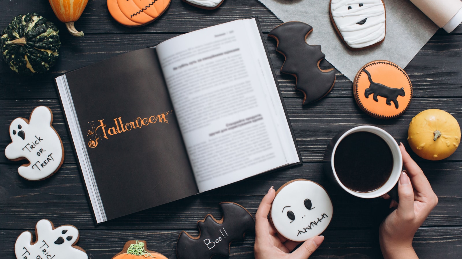 Girl holding cup of coffee, gingerbread for Halloween and reading horror book on a black wooden background.