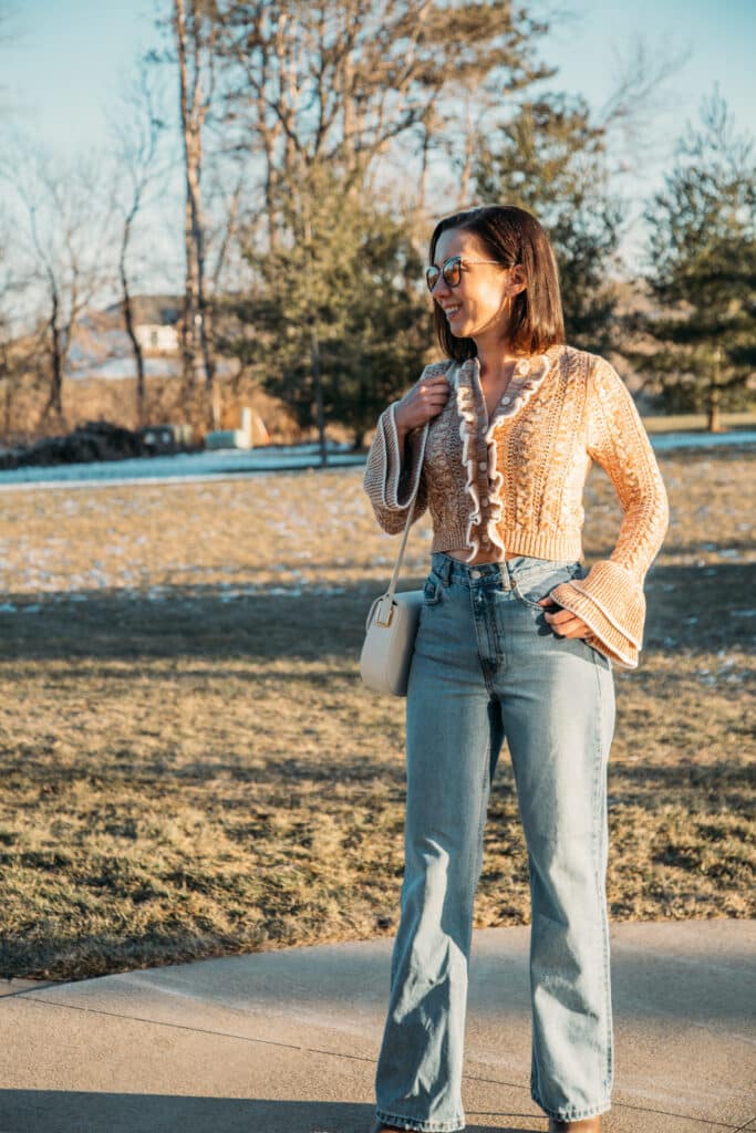 Lindsey wearing a cropped ruffle Revolve cardigan, wide leg jeans and ankle boots that have a spiky heel. She is standing on a cement side walk in the late afternoon with frozen grass and trees behind her.