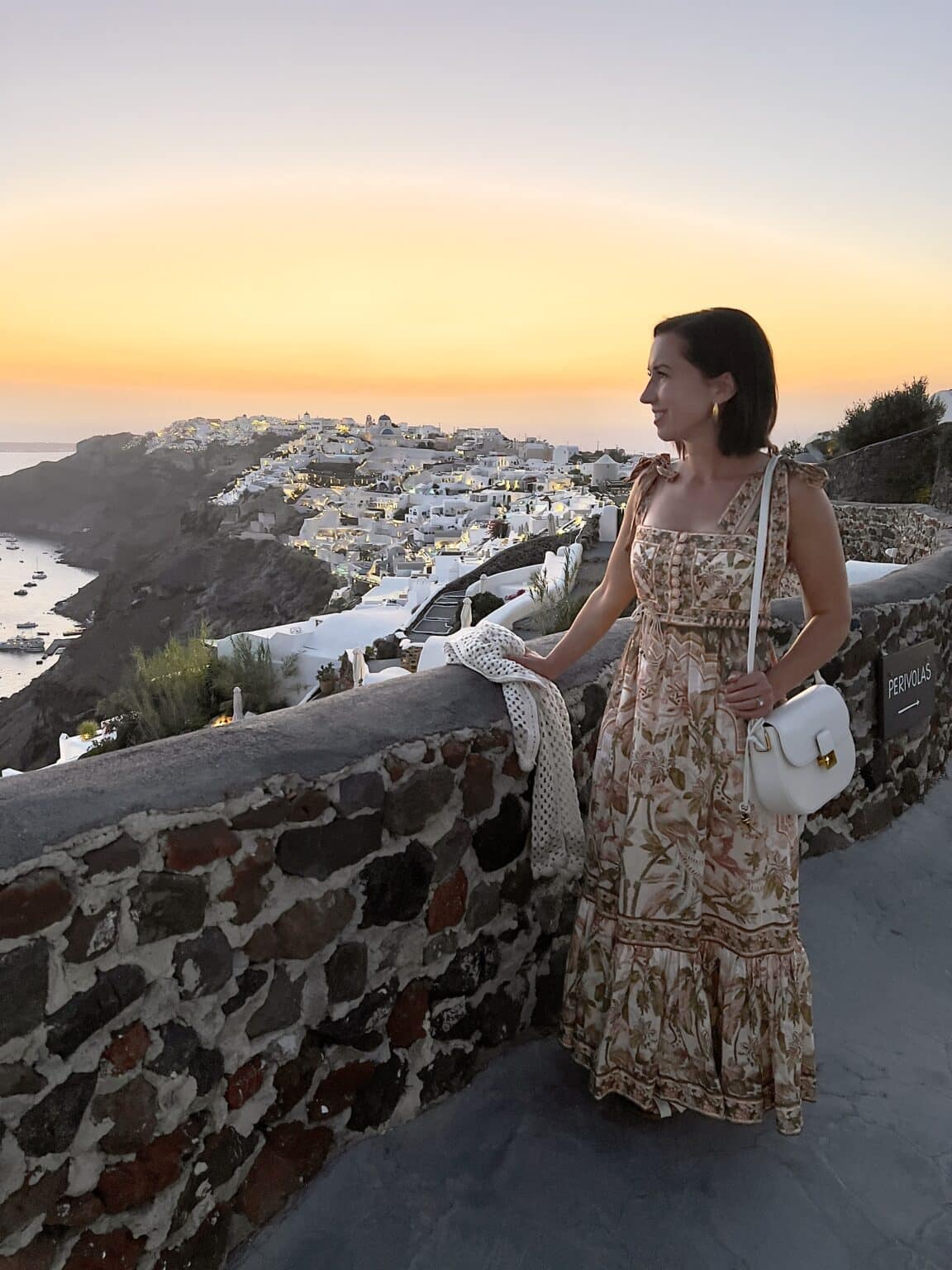 Lindsey of Have clothes, will travel wearing a floral Zimmermann dress with white Bottega Veneta bag looking at the sunset view of Santorini