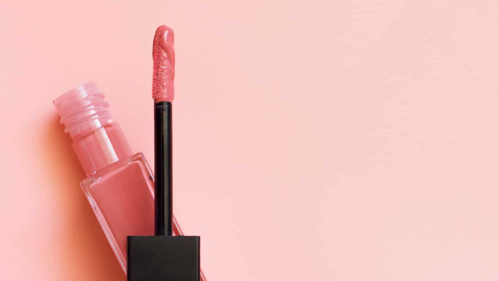 Liquid lipstick and applicator on pink color background. Open tube of lip gloss and wand brush with makeup product on pastel coral surface. Top view, flat lay, copy space