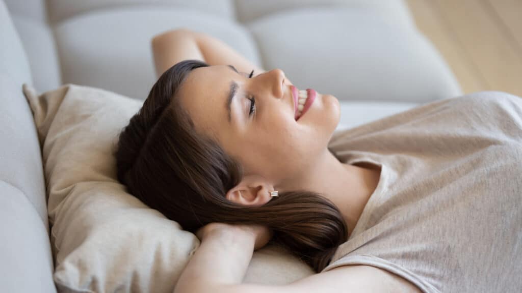 Happy relaxed woman lying on couch with eyes closed and enjoying peace and quiet of her own soundproof house. Good-looking young lady lounging on sofa cushion and smiling feeling content and fulfilled