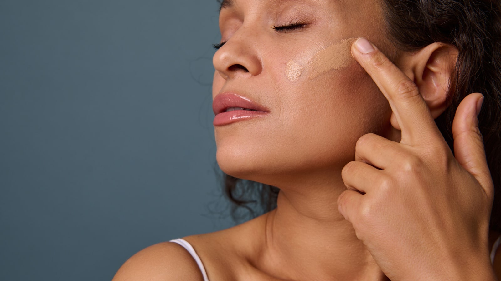 Close-up of attractive woman with her eyes closed applying liquid tonal foundation fluid on her face with her finger, posing against gray wall background with copy space. Make-up, skin care concept