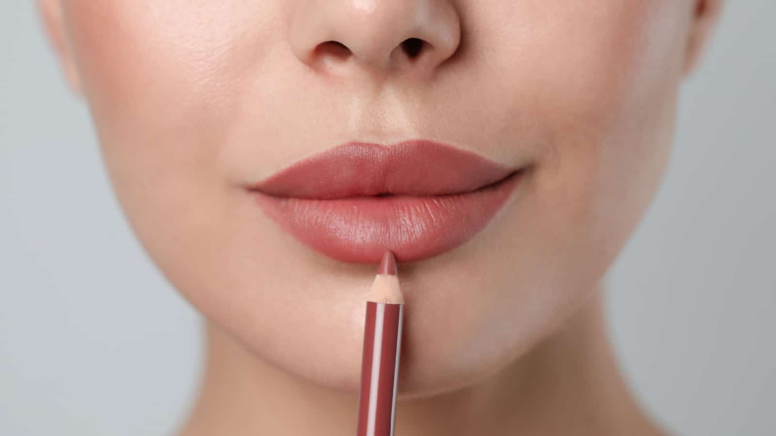 Young woman applying beautiful nude lip pencil on light grey background, closeup