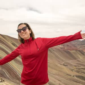 Lindsey wearing a read Qzip pullover from SCOTTeVEST with arms out stretched and Rainbow Mountain in Peru behind her.