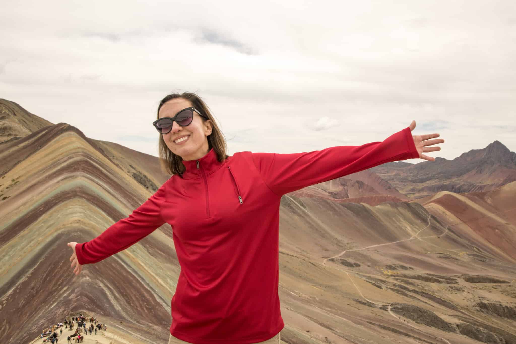 Lindsey wearing a read Qzip pullover from SCOTTeVEST with arms out stretched and Rainbow Mountain in Peru behind her.